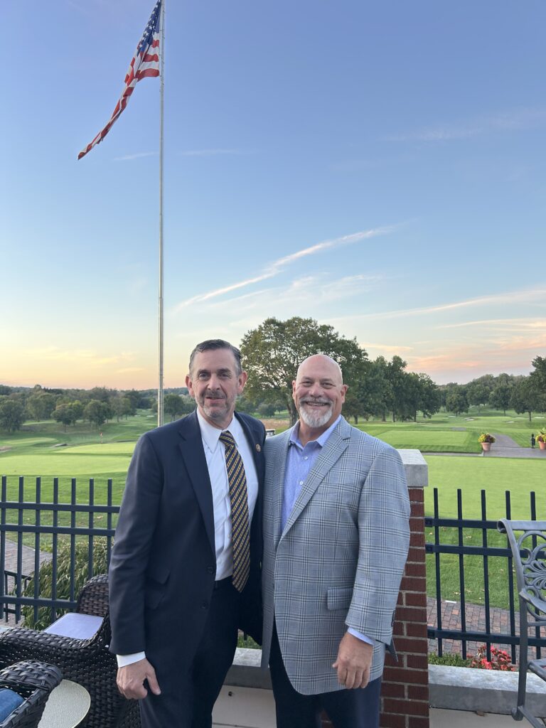 Two men smiling by a flagpole.